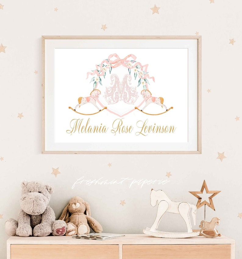 May include: A framed print featuring a watercolor design with a pink and gold color scheme. The artwork includes a floral wreath, two rocking horses, and the name "Melania Rose Levinson" in gold script. The print is displayed on a light-colored wall.