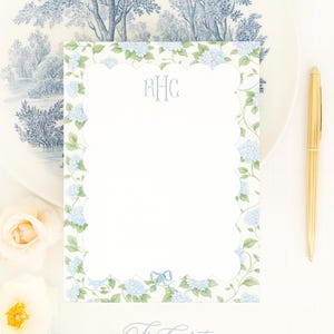 May include: A stationery set featuring a floral design with blue hydrangeas and green leaves. The set includes a notepad with a scalloped edge, a gold pen, and a decorative plate with a blue and white tree pattern. The notepad has the initials "RHC".
