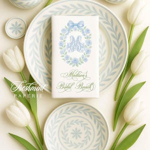 May include: An elegant bridal brunch table setting featuring white tulips, light blue floral patterned plates, and matching lidded jars. A napkin with a floral wreath and the text "Madison's Bridal Brunch" is centered on a plate.