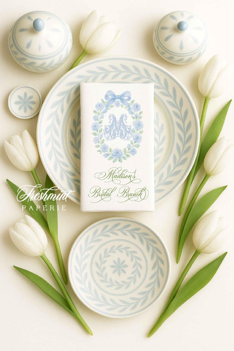 May include: An elegant bridal brunch table setting featuring white tulips, light blue floral patterned plates, and matching lidded jars. A napkin with a floral wreath and the text "Madison's Bridal Brunch" is centered on a plate.