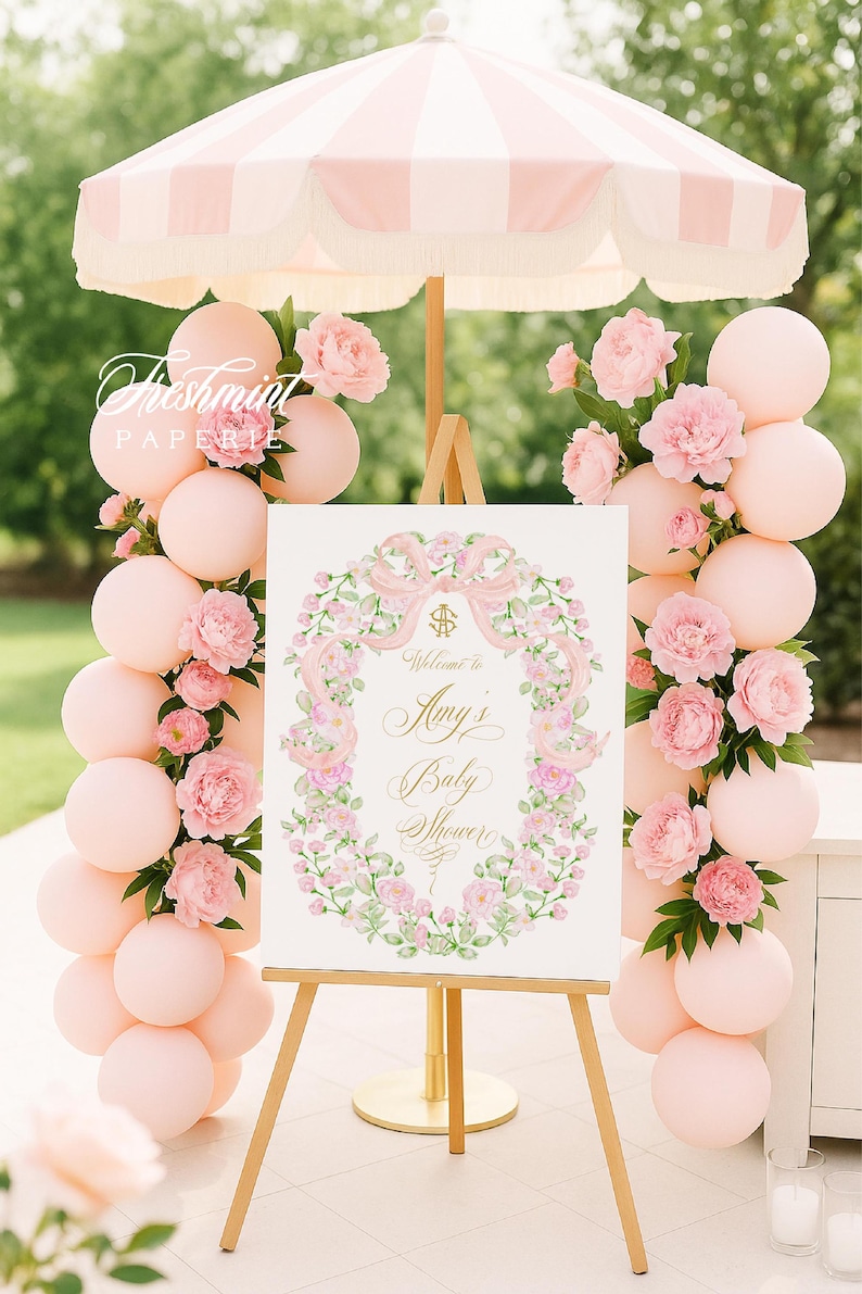 May include: A baby shower scene with a sign that reads "Welcome to Amy's Baby Shower." The sign is surrounded by a floral wreath and set on a wooden easel. Pink balloons and flowers decorate the scene, with a striped pink and white umbrella overhead.