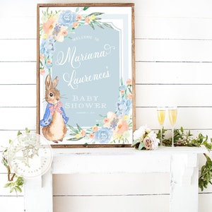 May include: A blue and white baby shower sign with a watercolour floral border and a cartoon rabbit. The sign says "Welcome to Mariana Laurence's Baby Shower".
