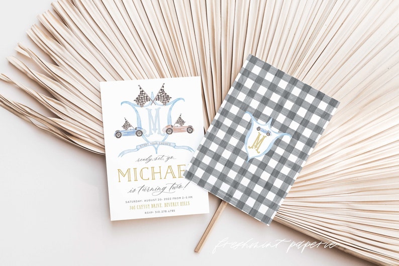 May include: A white invitation card with a blue and brown illustration of race cars and checkered flags. The text "Ready, set, go Michael is turning two!" is written in gold lettering. The invitation includes the date, time, and address for the event.