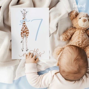 K&ouml;nnte beinhalten: Ein Baby h&auml;lt eine wei&szlig;e Karte mit einer blauen Zahl Sieben und einer Giraffenillustration. Die Karte sagt "7 Monate".