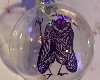 May include: Clear glass Christmas ornament with a dark purple moth design. The moth has red eyes and is holding a staff. The ornament has a silver metal cap and a wire for hanging. The moth design is a linocut style.
