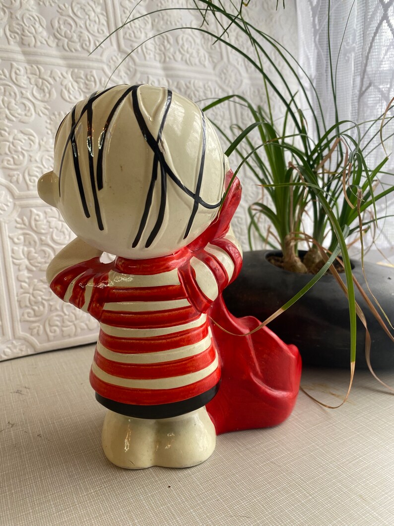 May include: A ceramic figurine of a child holding a red guitar. The child is wearing a red and white striped shirt and black trousers. The figurine is painted in a whimsical style.