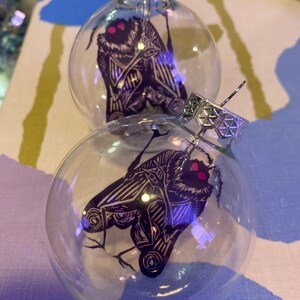 May include: Two clear glass Christmas ornaments with silver tops. Each ornament features a black and white moth design with pink eyes. The ornaments are displayed on a patterned surface.