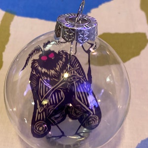 May include: Clear glass Christmas ornament with a silver metal cap. The ornament features a dark blue moth illustration with pink eyes and gold accents. The moth design is visible inside the clear glass sphere.