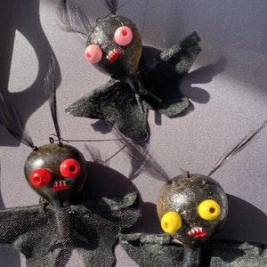 May include: Three black felt bat ornaments with different colored eyes. The bats have black felt wings and black felt bodies. The eyes are red, pink, and yellow.