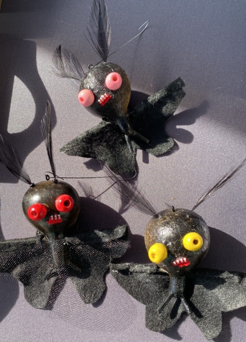 May include: Three black felt bat ornaments with different colored eyes. The bats have black felt wings and black felt bodies. The eyes are red, pink, and yellow.