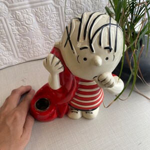 May include: Ceramic figurine of a cartoon boy with black hair, wearing a red and white striped shirt and red trousers. The figurine has a hole in the back, suggesting it may be a piggy bank or a planter.