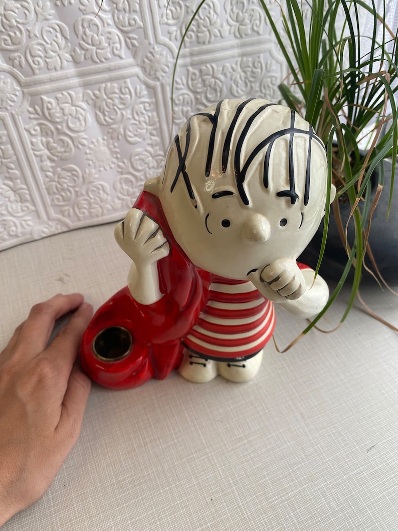 May include: Ceramic figurine of a cartoon boy with black hair, wearing a red and white striped shirt and red trousers. The figurine has a hole in the back, suggesting it may be a piggy bank or a planter.
