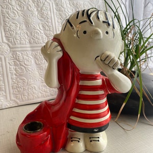 May include: A ceramic figurine of a boy wearing a red and white striped shirt and a red cape. The figurine has a candle holder on the side.