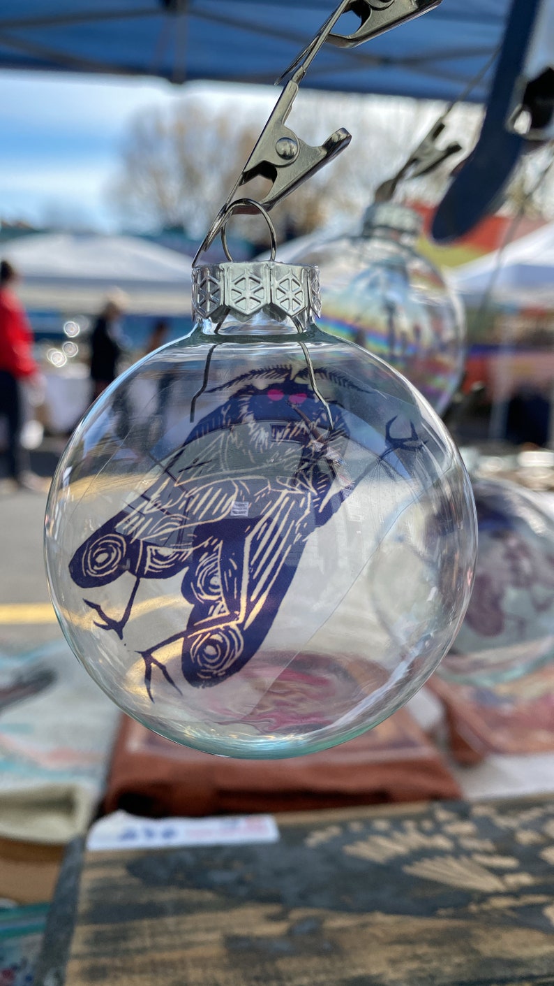 May include: Clear glass Christmas ornament with a detailed blue and white moth illustration. The ornament is suspended by a silver clip. The moth design features intricate wing patterns and is visible through the transparent glass.