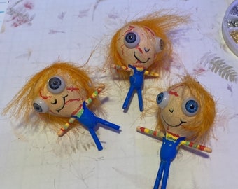 May include: Three handmade dolls with orange yarn hair, blue overalls, and large, white eyes. The dolls have red painted lines on their faces and are smiling.