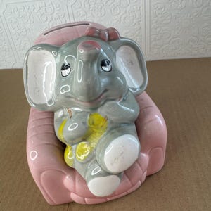 May include: A ceramic elephant-shaped piggy bank in shades of gray, pink, and yellow. The elephant has large ears, a trunk, and a red bow. It is cradled in a pink hand-shaped base, with a slot for coins on top.