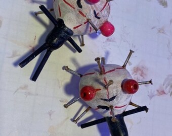 May include: Two small, white, round, doll-like figures with red painted eyes and mouths. Each figure has a black stick figure body and is covered in small silver nails.