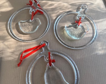 May include: Three clear glass ornaments with red ribbons. Two ornaments feature a bird design, and one has a bell shape. Each ornament is suspended within a circular frame, creating a delicate and festive appearance.