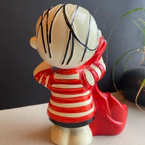 May include: A ceramic figurine of a boy with black hair, wearing a red and white striped shirt and holding a red bag. The figurine is standing on a white surface.