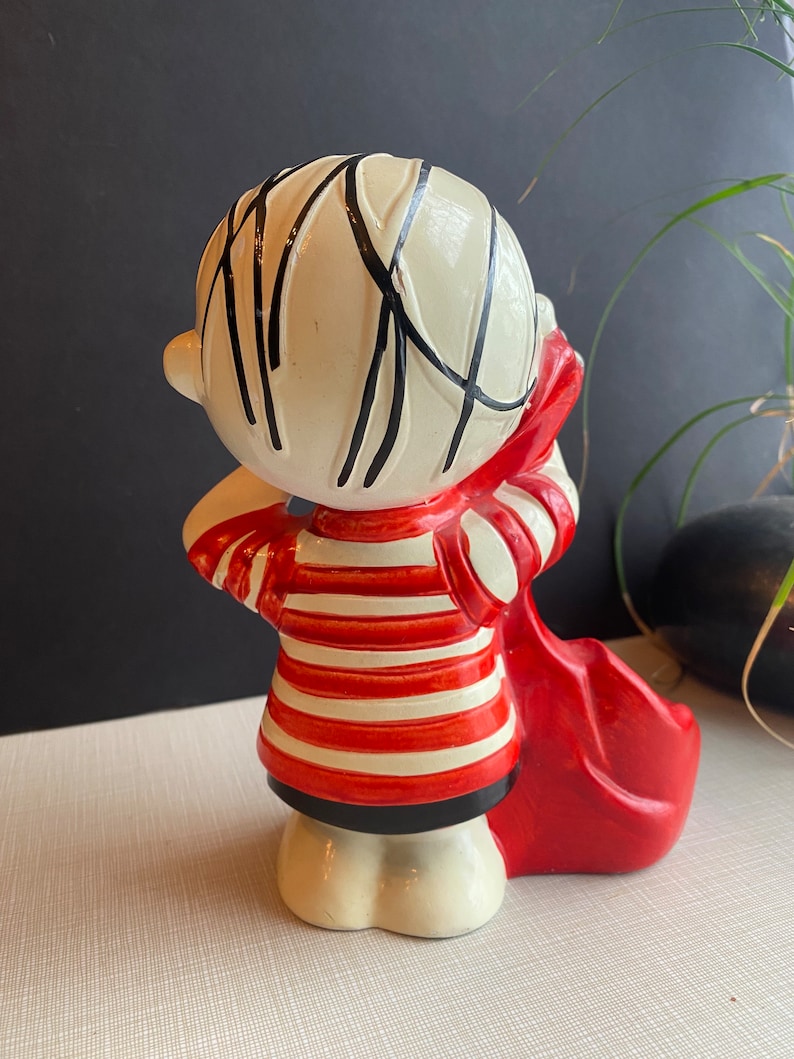 May include: A ceramic figurine of a boy with black hair, wearing a red and white striped shirt and holding a red bag. The figurine is standing on a white surface.