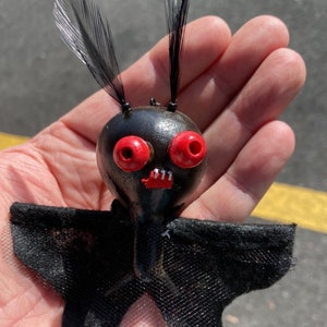 May include: A black bat-shaped ornament with red eyes and a toothy grin. The bat has black feathers for ears and a black tulle skirt.