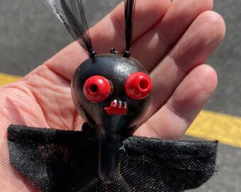 May include: A black bat-shaped ornament with red eyes and a toothy grin. The bat has black feathers for ears and a black tulle skirt.
