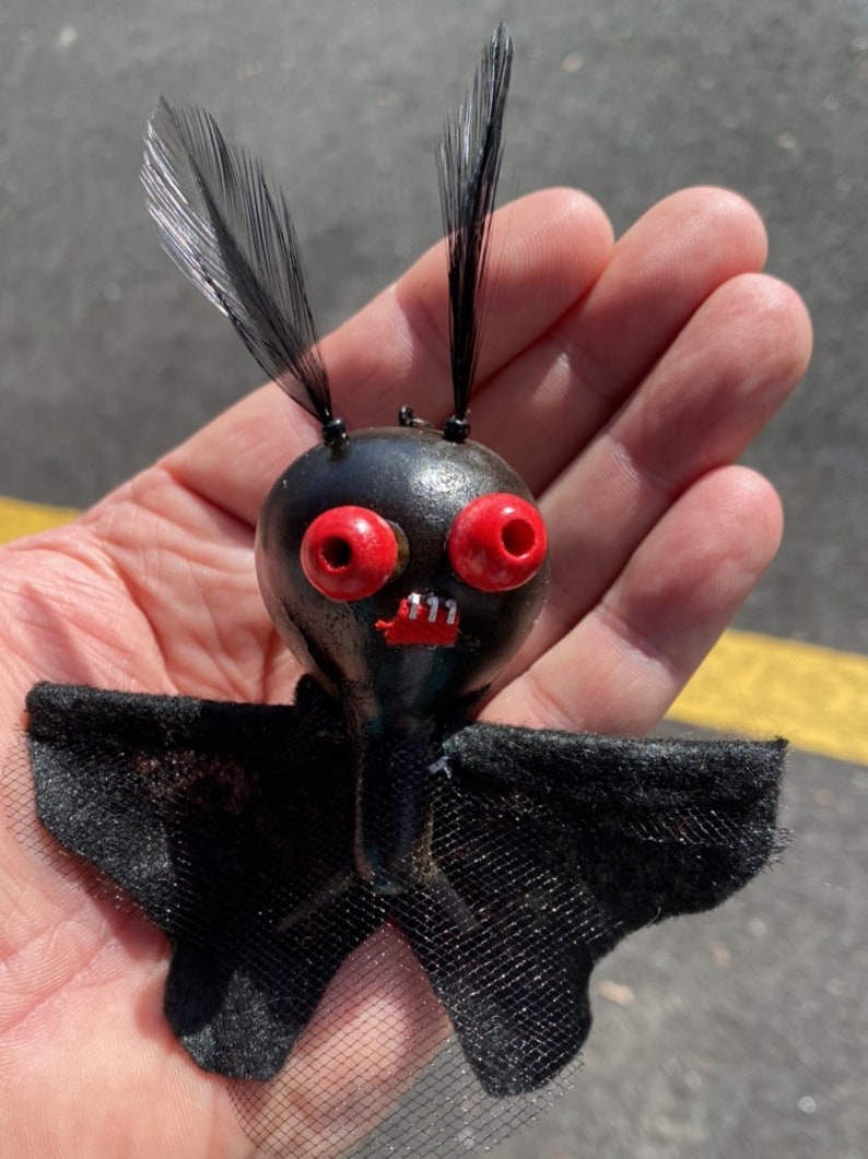 May include: A black bat-shaped ornament with red eyes and a toothy grin. The bat has black feathers for ears and a black tulle skirt.
