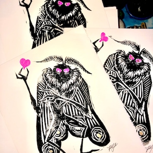 May include: Three black and white linocut prints of a moth-like creature with pink hearts. The creature has large eyes and a striped body. The prints are signed "Rye".