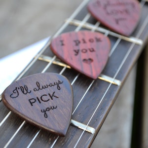 May include: Three wooden guitar picks with different messages engraved on them. The picks are arranged on a guitar neck. The messages are "I Pick You", "I'll Always Pick You", and "Always Love You".