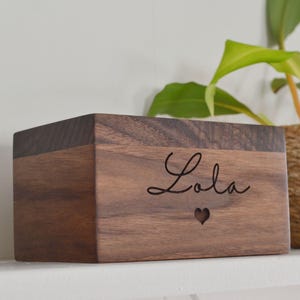 May include: A wooden box with the name "Lola" engraved on the front. The box is made of dark walnut wood and has a heart shape carved into the front.