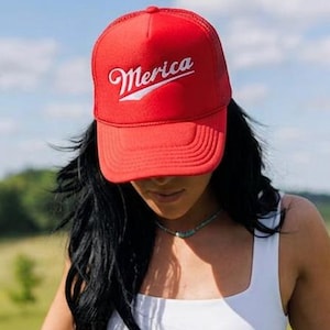 Könnte beinhalten: Eine leuchtend rote Trucker-Cap mit dem in weißer Schreibschrift aufgestickten Wort Merica auf der Vorderseite, kombiniert mit einem weißen ärmellosen Top und einer blauen Jeans mit horizontalen Schlitzen, die mit silbernen Strasssteinen verziert sind.