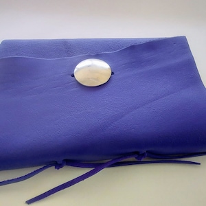 May include: A handmade, royal blue leather pouch with a large silver button closure. The pouch is rectangular with a folded top and features a leather tie closure. The leather has a textured appearance.