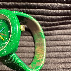 May include: A green watch with a white speckled face and a green expandable band. The watch face has a white ring around the edge with the words "Swiss Made" printed on it.