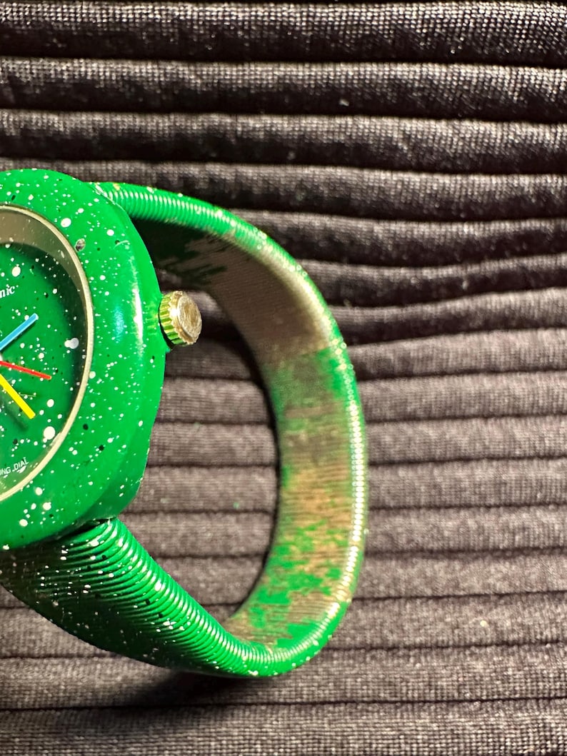 May include: A green watch with a white speckled face and a green expandable band. The watch face has a white ring around the edge with the words "Swiss Made" printed on it.