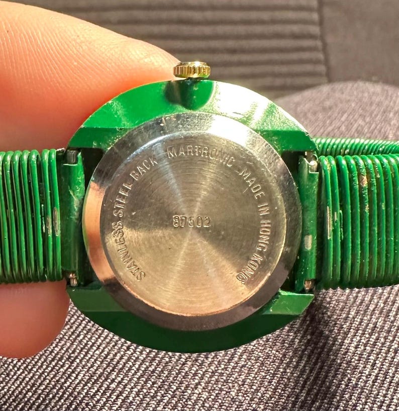 May include: A green wristwatch with a stainless steel back. The watch face is silver and has the text "Marlboro Made in Hong Kong" and "Stainless Steel Back".