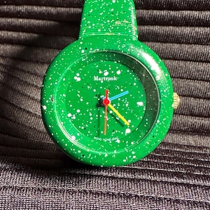 May include: A green wristwatch with white speckles on the face and band. The watch face has a white background with black text that reads "Martronic" and "HONG KONG DIAL". The watch hands are red, blue, and yellow.