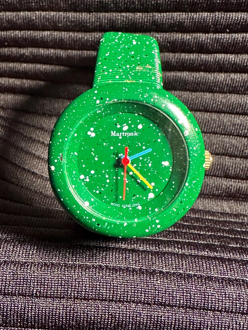 May include: A green wristwatch with white speckles on the face and band. The watch face has a white background with black text that reads "Martronic" and "HONG KONG DIAL". The watch hands are red, blue, and yellow.