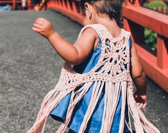 Op de afbeelding: Een lichtroze gehaakt vest met franjes, gedragen over een blauwe spijkerjurk. Het vest heeft een open ontwerp op de rug. Het kind loopt op een grijs oppervlak met een rode brug op de achtergrond.