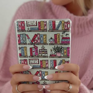 May include: A clear phone case featuring a colorful bookshelf design. The illustration includes books, candles, and potted plants. The words "GOOD GIRL" are visible on the case. The case is held in front of a pink sweater.
