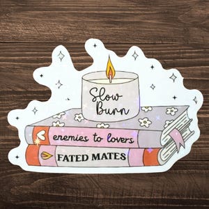 May include: A sticker featuring a stack of books with the titles "enemies to lovers" and "FATED MATES". A lit candle with the text "Slow Burn" sits atop the books. The sticker has a white outline with star and diamond accents.