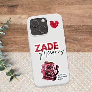 K&ouml;nnte beinhalten: Wei&szlig;es Handycase mit einem roten Glitzer-Herz-Aufkleber, dem Namen "ZADE Meadows" in roter und schwarzer Schrift und einem dunkelroten Rosenaufkleber. Der Text "MEADOWS, BABY. OUR LAST NAME IS MEADOWS" ist ebenfalls vorhanden.