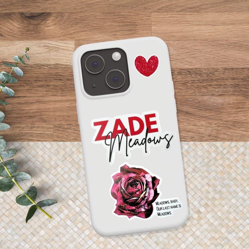 K&ouml;nnte beinhalten: Wei&szlig;es Handycase mit einem roten Glitzer-Herz-Aufkleber, dem Namen "ZADE Meadows" in roter und schwarzer Schrift und einem dunkelroten Rosenaufkleber. Der Text "MEADOWS, BABY. OUR LAST NAME IS MEADOWS" ist ebenfalls vorhanden.