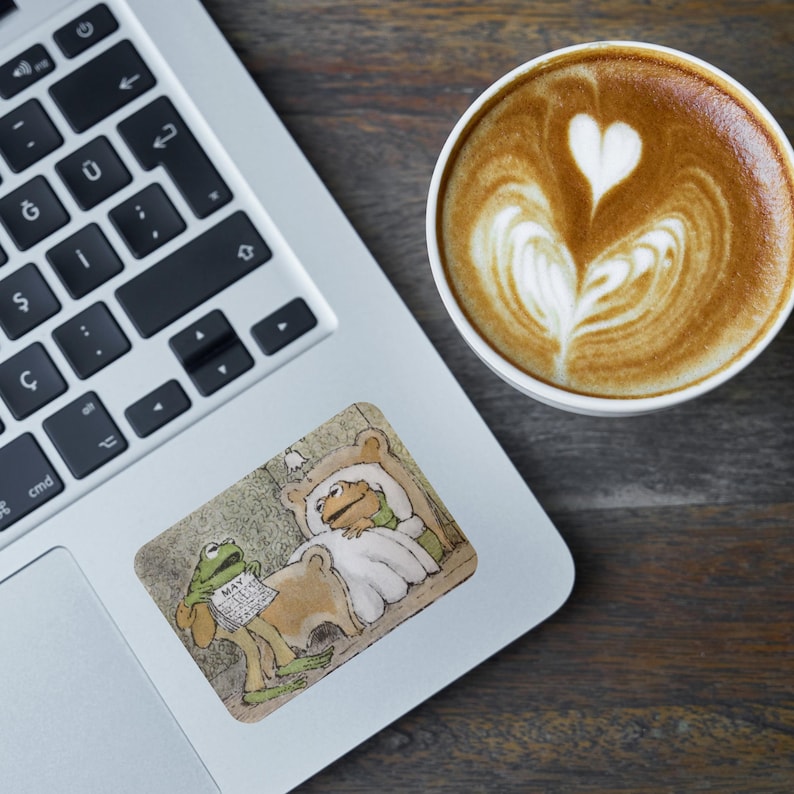 Puede incluir: Una pegatina para port&aacute;til con una rana de dibujos animados leyendo un peri&oacute;dico con la palabra "MAY" visible, mientras que otra rana descansa en la cama. Una taza de caf&eacute; con arte latte est&aacute; al lado del port&aacute;til sobre una superficie de madera.