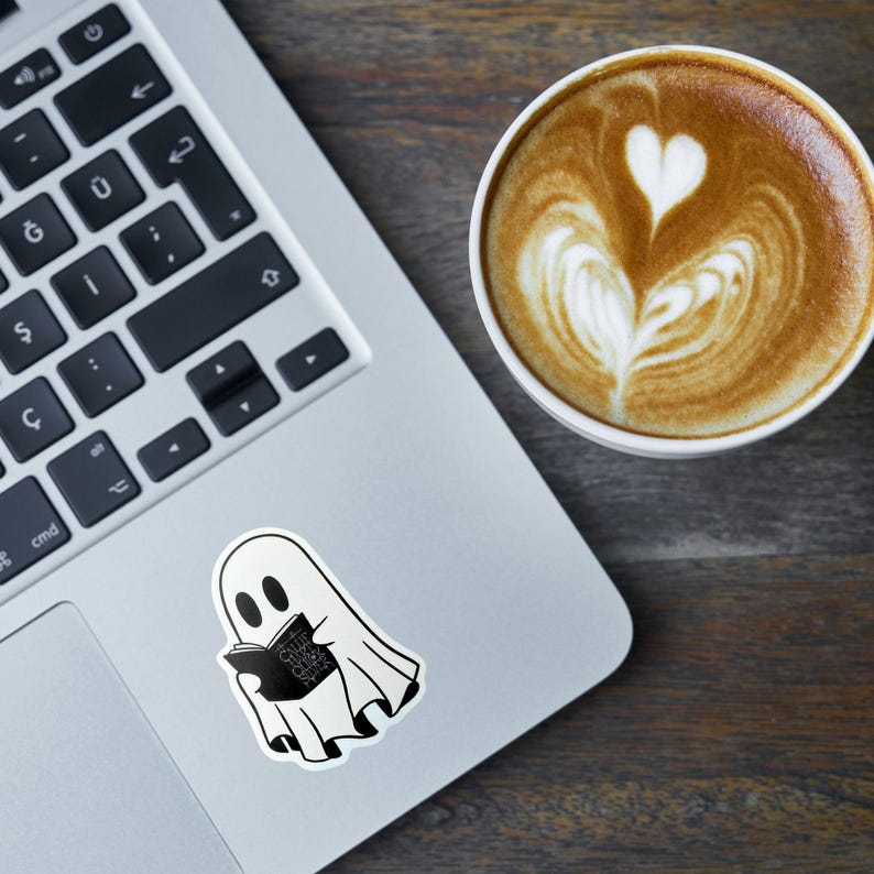May include: A white ghost sticker reading a book with the text "Spooky Stories" in black. The sticker is on a laptop next to a cup of coffee with latte art.