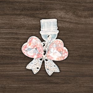 May include: A clear bottle stopper with a bow-shaped design. The bow features two heart-shaped pink crystals and a lower section with small, clear crystals. The stopper is made of clear plastic and has a silver-colored metal stem.