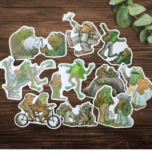 Puede incluir: Una colecci&oacute;n de pegatinas de ranas caprichosas en varias poses y atuendos. Las ranas est&aacute;n representadas en un estilo de dibujos animados, algunas sosteniendo teteras, leyendo libros o montando en bicicleta. Las pegatinas tienen un borde blanco.