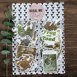 Puede incluir: Un conjunto de pegatinas de Frog and Toad en una bolsa transparente con un patr&oacute;n de tri&aacute;ngulos en blanco y negro. Las pegatinas presentan ilustraciones de los personajes, con el texto "Frog and Toad" en verde.
