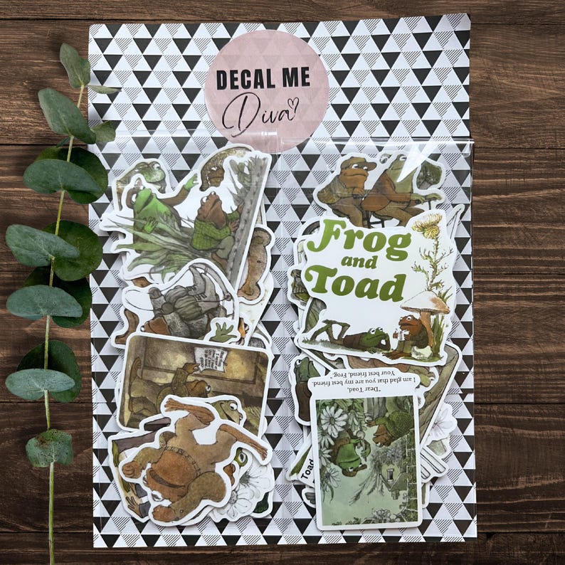 Puede incluir: Un conjunto de pegatinas de Frog and Toad en una bolsa transparente con un patr&oacute;n de tri&aacute;ngulos en blanco y negro. Las pegatinas presentan ilustraciones de los personajes, con el texto "Frog and Toad" en verde.