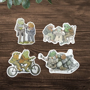 Puede incluir: Conjunto de cuatro pegatinas caprichosas con ranas antropom&oacute;rficas en varias escenas. Una pegatina muestra dos ranas con ropa formal, otra representa ranas en una mesa, una tercera muestra ranas en bicicleta y la &uacute;ltima muestra ranas esquiando.