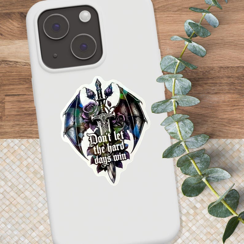 K&ouml;nnte beinhalten: Eine wei&szlig;e Smartphone-H&uuml;lle mit einem Aufkleber, der ein zentrales silbernes Schwert, dunkelviolette Rosen und drachenartige Fl&uuml;gel in Blau- und Gr&uuml;nt&ouml;nen zeigt. Der Text "Don't let the hard days win" ist in einer gotischen Schrift gedruckt.